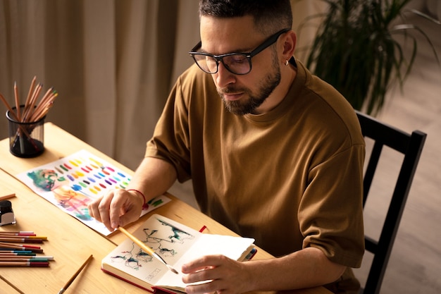 Retrato de joven artista masculino blanco caucásico concentrado de pelo oscuro con barba en gafas, camiseta de color caqui. sentado en un estudio en una mesa de madera, sosteniendo un pincel en la mano. Bricolaje.