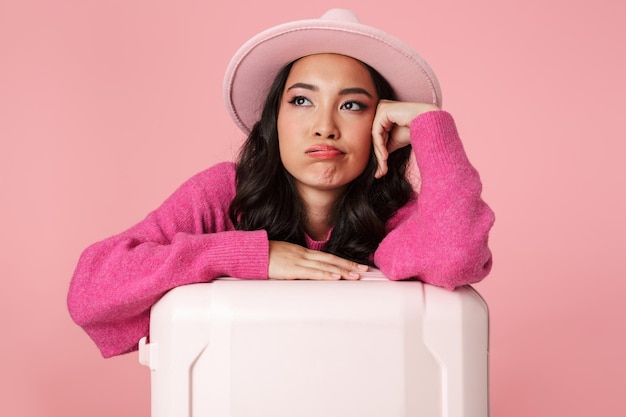 Retrato de joven asiática aburrida con sombrero sentado con maleta de viaje aislado en rosa