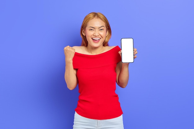 Retrato de una joven asiática alegre que muestra un teléfono móvil con pantalla en blanco y hace un gesto de éxito aislado en un fondo morado