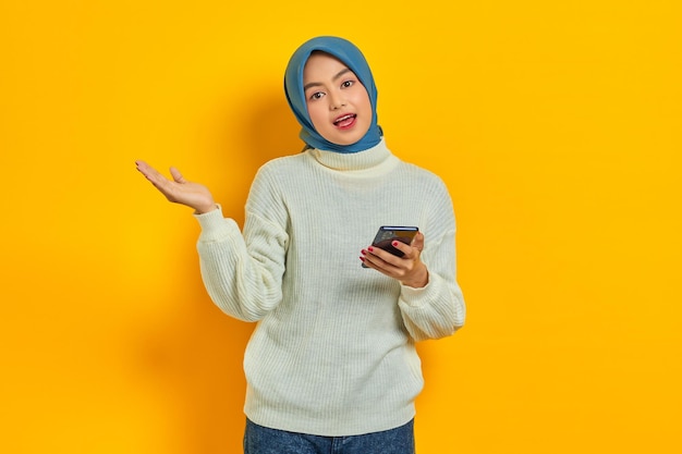 Retrato de una joven asiática alegre con suéter blanco y hiyab sosteniendo un teléfono móvil aislado sobre un fondo amarillo Concepto religioso del islam de la gente