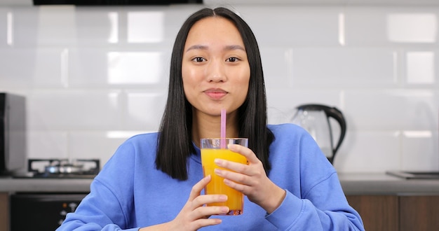 Retrato de una joven asiática bebiendo jugo de naranja una atractiva chica morena multirracial está sentada...
