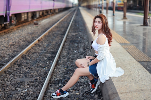 Foto retrato de joven asiática, cabello largo con vestido blanco sentado y mirando a la cámara mientras espera en la estación de tren con cara de belleza