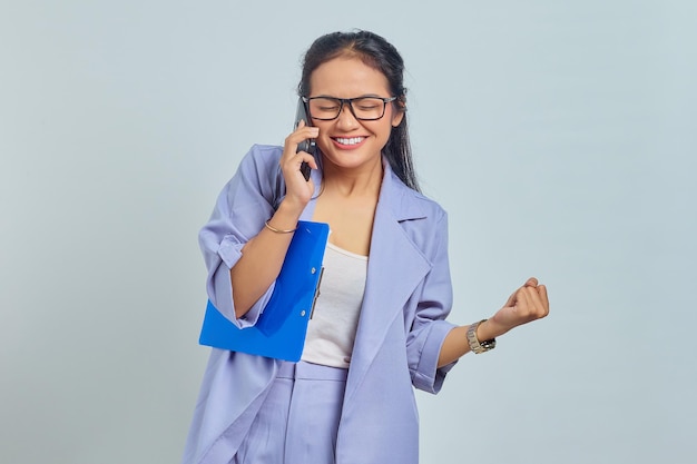 Retrato de una joven asiática emocionada hablando por teléfono móvil y contenta de lograr la victoria aislada en un fondo morado