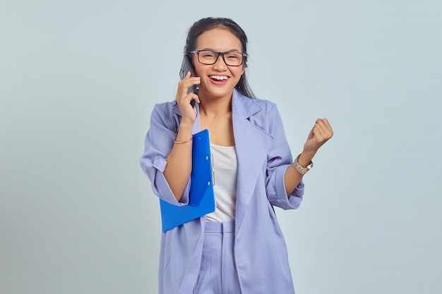 Retrato de una joven asiática emocionada hablando por teléfono móvil y contenta de lograr la victoria aislada en un fondo morado