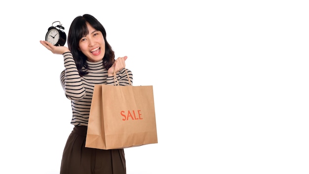 Retrato de una joven asiática feliz con una camisa de suéter sosteniendo un despertador y un papel de compras aislado de fondo blanco