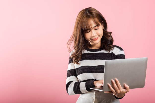 Retrato de una joven asiática feliz, hermosa, confiada, con la cara sonriente, sosteniendo usando una computadora portátil, mirando a la cámara, grabado en un estudio aislado sobre un fondo rosa con espacio para copiar.