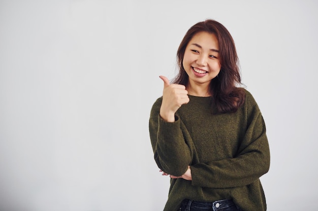 Retrato de una joven asiática feliz que muestra el pulgar hacia arriba en el interior del estudio con fondo blanco.