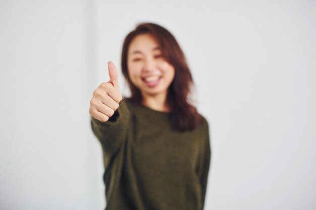 Retrato de una joven asiática feliz que muestra el pulgar hacia arriba en el interior del estudio con fondo blanco.