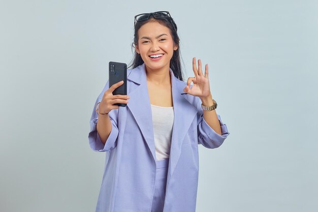 Retrato de una joven asiática feliz usando un teléfono móvil y gesticulando con un signo de ok aislado de fondo blanco
