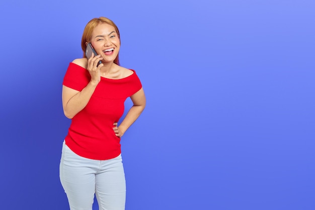 Retrato de una joven asiática feliz vestida de rojo regocijándose mientras habla por teléfono móvil aislada de fondo morado