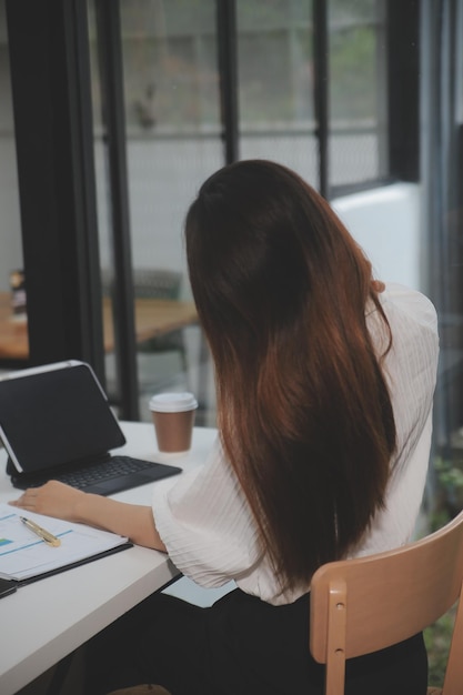 Retrato de una joven asiática independiente está trabajando en su trabajo en una tableta de computadora en una oficina moderna. Haciendo un informe de análisis contable. Datos de inversión inmobiliaria. Concepto de sistemas financieros y fiscales.