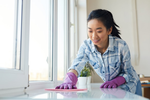 Retrato de joven asiática lavando ventanas y sonriendo felizmente mientras disfruta de la limpieza de primavera