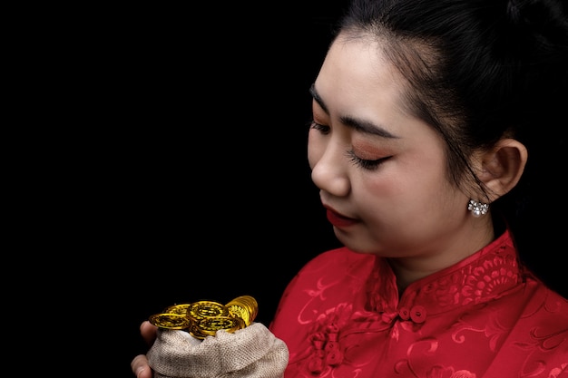 Retrato joven asiática, mujer vestido rojo cheongsam tradicional sosteniendo una moneda de oro en un saco en el fondo negro