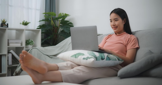 Retrato de una joven asiática relajándose en casa sentada en un cómodo sofá usando una computadora portátil hablando con amigos en redes sociales haciendo compras de trabajo remotas en línea