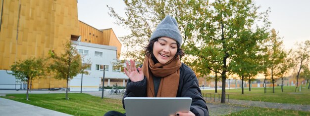Retrato de una joven asiática sentada con ropa abrigada en el parque ondas de la mano en videoconferencias de tabletas al aire libre