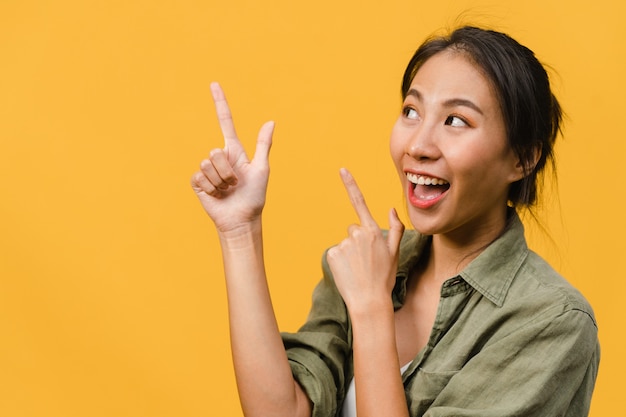 Retrato de joven asiática sonriendo con expresión alegre, muestra algo sorprendente en el espacio en blanco en ropa casual y de pie aislado sobre una pared amarilla. Concepto de expresión facial.