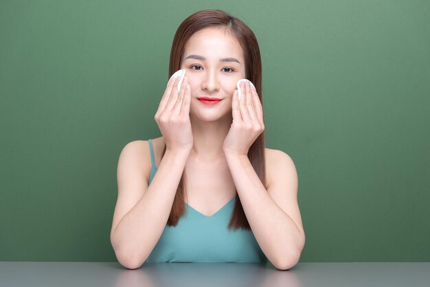 Retrato de una joven asiática sonriente limpiándose la cara con una almohadilla de algodón aislada sobre el fondo verde