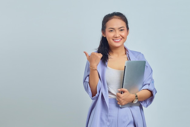 Foto retrato de una joven asiática sonriente sosteniendo un portátil y mostrando un gesto con el pulgar hacia arriba aislado de fondo blanco