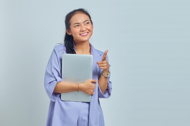 Retrato de una joven asiática sonriente sosteniendo un portátil y señalando con el dedo a la cámara aislada de fondo blanco