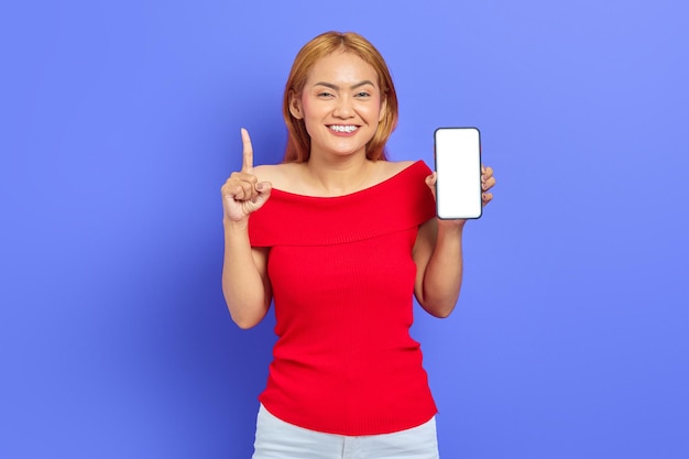 Retrato de una joven asiática sonriente sosteniendo un teléfono inteligente y señalando con el dedo el espacio de copia aislado en un fondo morado