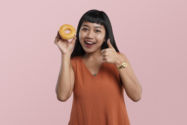 Retrato de una joven asiática sosteniendo donuts con los pulgares hacia arriba, aislada sobre fondo rosa