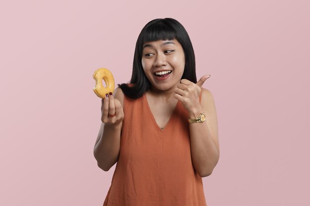 Retrato de una joven asiática sosteniendo donuts con los pulgares hacia arriba, aislada sobre fondo rosa