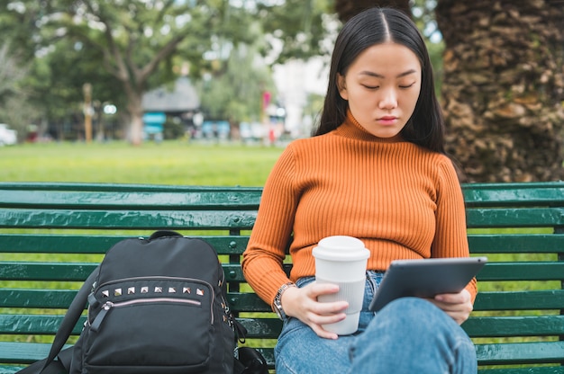 Retrato de joven asiática con su tableta digital mientras sostiene una taza de café en el parque al aire libre.