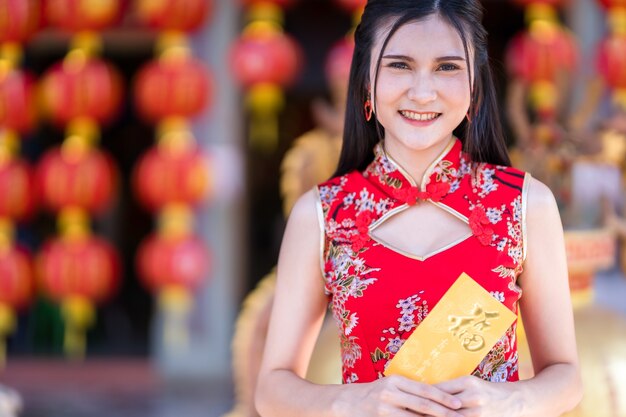 Retrato de joven asiática vistiendo cheongsam chino tradicional rojo, sosteniendo sobres amarillos con el texto chino Bendiciones escrito en él Es una buena suerte para el Festival del Año Nuevo Chino