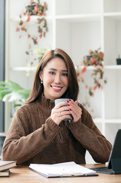 Retrato de joven asiática vistiendo suéter de lana sosteniendo la taza, sonriendo y mirando a la cámara mientras está sentado en el escritorio de oficina. Vista vertical.