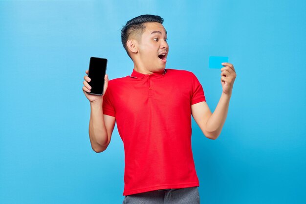 Retrato de un joven asiático alegre que muestra un teléfono móvil con pantalla en blanco y una tarjeta de crédito aislada de fondo azul