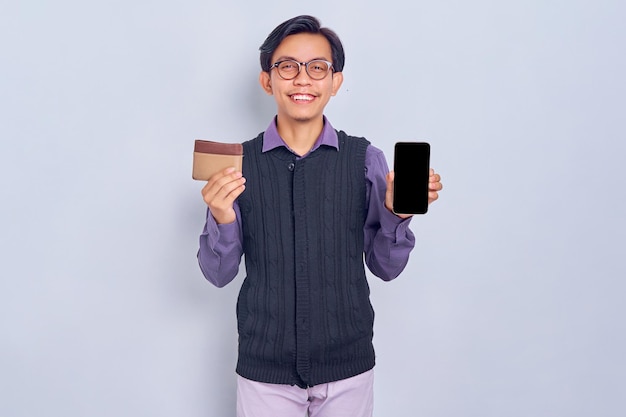 Retrato de un joven asiático alegre que usa una camisa que muestra un teléfono móvil con pantalla en blanco y una billetera marrón aislada de fondo blanco