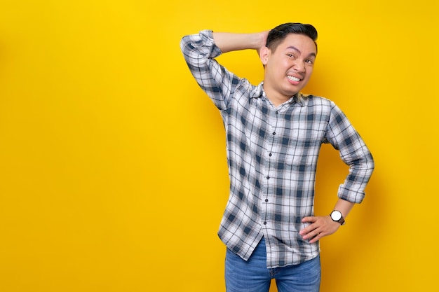 Retrato de un joven asiático confundido con camisa a cuadros con la mano en la cabeza rascándose la cintura mientras mira a un lado en el área del espacio de trabajo simulado aislado en el concepto de estilo de vida de la gente de fondo amarillo