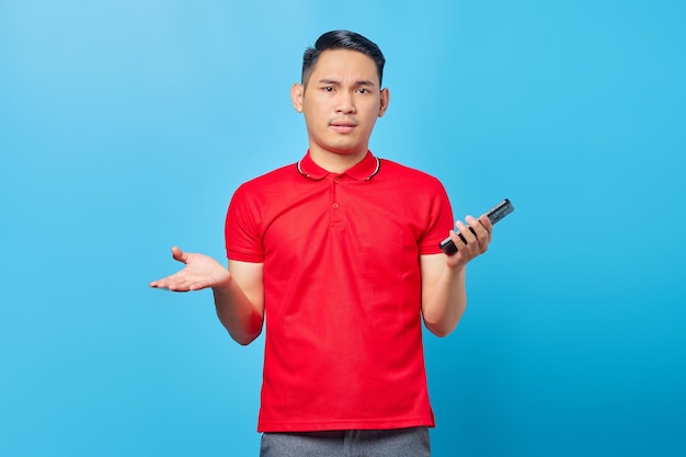 Retrato de un joven asiático confundido usando un teléfono móvil y mirando una cámara aislada de fondo azul