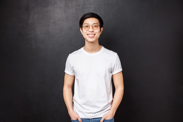 Retrato de un joven asiático con gafas de pie en camisa casual blanca sobre, sonriendo amistosamente, expresa feliz emoción entusiasta, colgando con compañeros después de la universidad