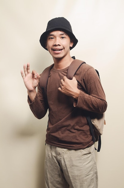 Retrato de un joven asiático haciendo un buen gesto llevando una mochila para viajar aislado en el fondo