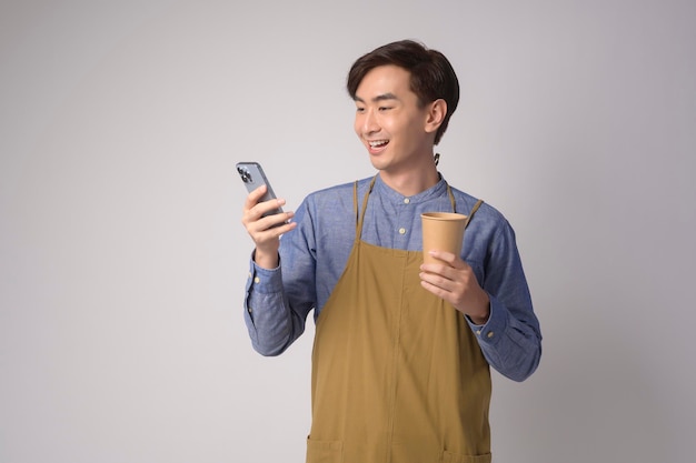 retrato, de, joven, asiático, hombre, llevando, delantal, tenencia, teléfono inteligente, y, taza de papel, encima, fondo blanco