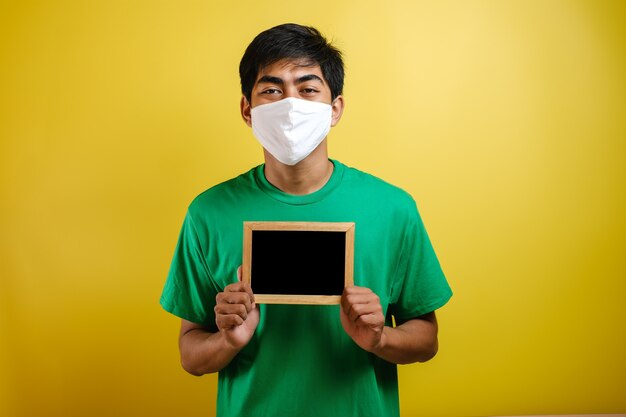 Retrato de joven asiático con máscara protectora contra el coronavirus, sosteniendo una pequeña pizarra sobre fondo amarillo