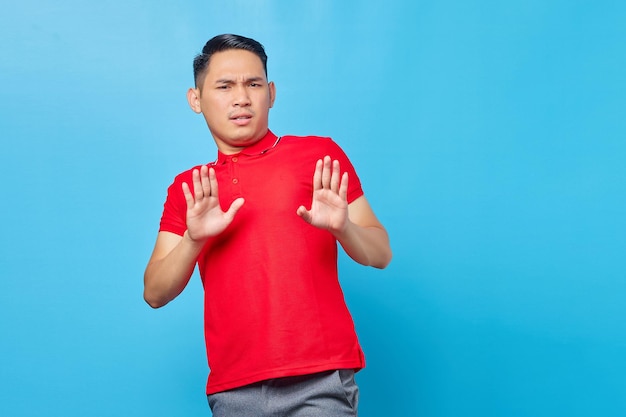 Retrato de un joven asiático molesto que cruza las manos hace un gesto de parada, demuestra rechazo aislado en el fondo azul