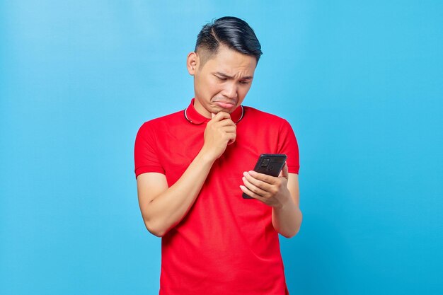 Retrato de un joven asiático pensativo mirando el teléfono móvil, leyendo noticias falsas aisladas de fondo azul