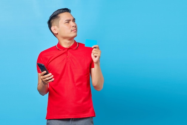 Retrato de un joven asiático pensativo que sostiene un teléfono móvil y una tarjeta de crédito aislado de fondo azul