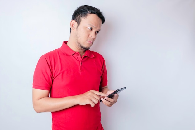 Retrato de un joven asiático pensativo que usa una camiseta roja mirando a un lado mientras sostiene un teléfono inteligente