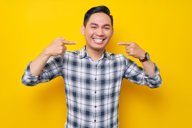 Retrato de un joven asiático sonriente con camisa a cuadros señalando con el dedo los dientes sonrisa dentuda blanca aislada en el concepto de estilo de vida de la gente de fondo amarillo