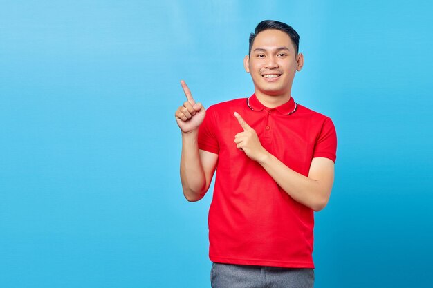 Retrato de un joven asiático sonriente con camisa roja de pie señalando con el dedo en el espacio de copia aislado de fondo azul