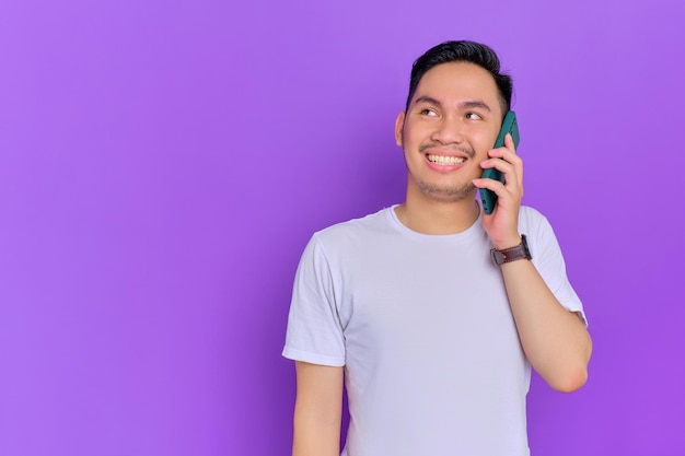 Retrato de un joven asiático sonriente con camiseta blanca hablando por teléfono móvil con espacio para copiar aislado de fondo morado