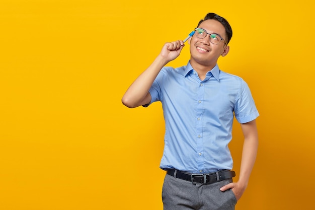 Retrato de un joven asiático sonriente con gafas que tiene una idea creativa y sostiene un bolígrafo aislado en un concepto de empresario y emprendedor de fondo amarillo