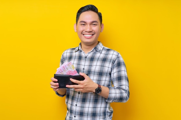 Retrato de un joven asiático sonriente con pantalones a cuadros sosteniendo una billetera llena de dinero en efectivo en billetes de rupias en la mano aislado en el concepto de estilo de vida de la gente de fondo amarillo