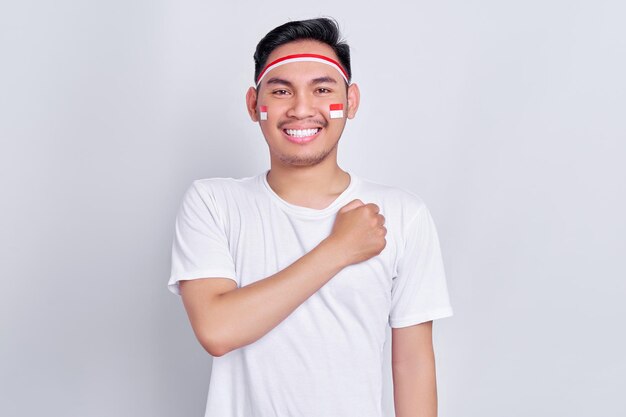 Retrato de un joven asiático sonriente que muestra un gesto de respeto con la mano en el pecho mientras celebra el día de la independencia de Indonesia el 17 de agosto aislado de fondo blanco