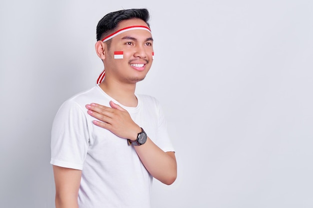 Retrato de un joven asiático sonriente que muestra un gesto de respeto con la mano en el pecho mientras celebra el día de la independencia de Indonesia el 17 de agosto aislado de fondo blanco