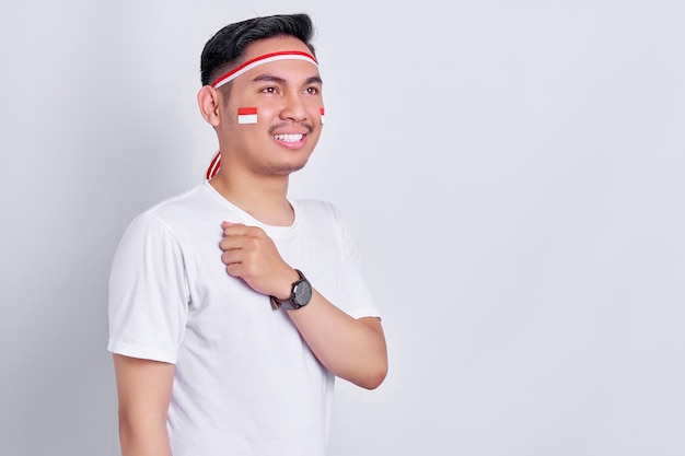 Retrato de un joven asiático sonriente que muestra un gesto de respeto con la mano en el pecho mientras celebra el día de la independencia de Indonesia el 17 de agosto aislado de fondo blanco