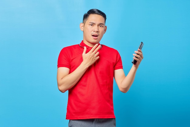 Retrato de un joven asiático sorprendido usando un teléfono móvil y mirando una cámara aislada de fondo azul